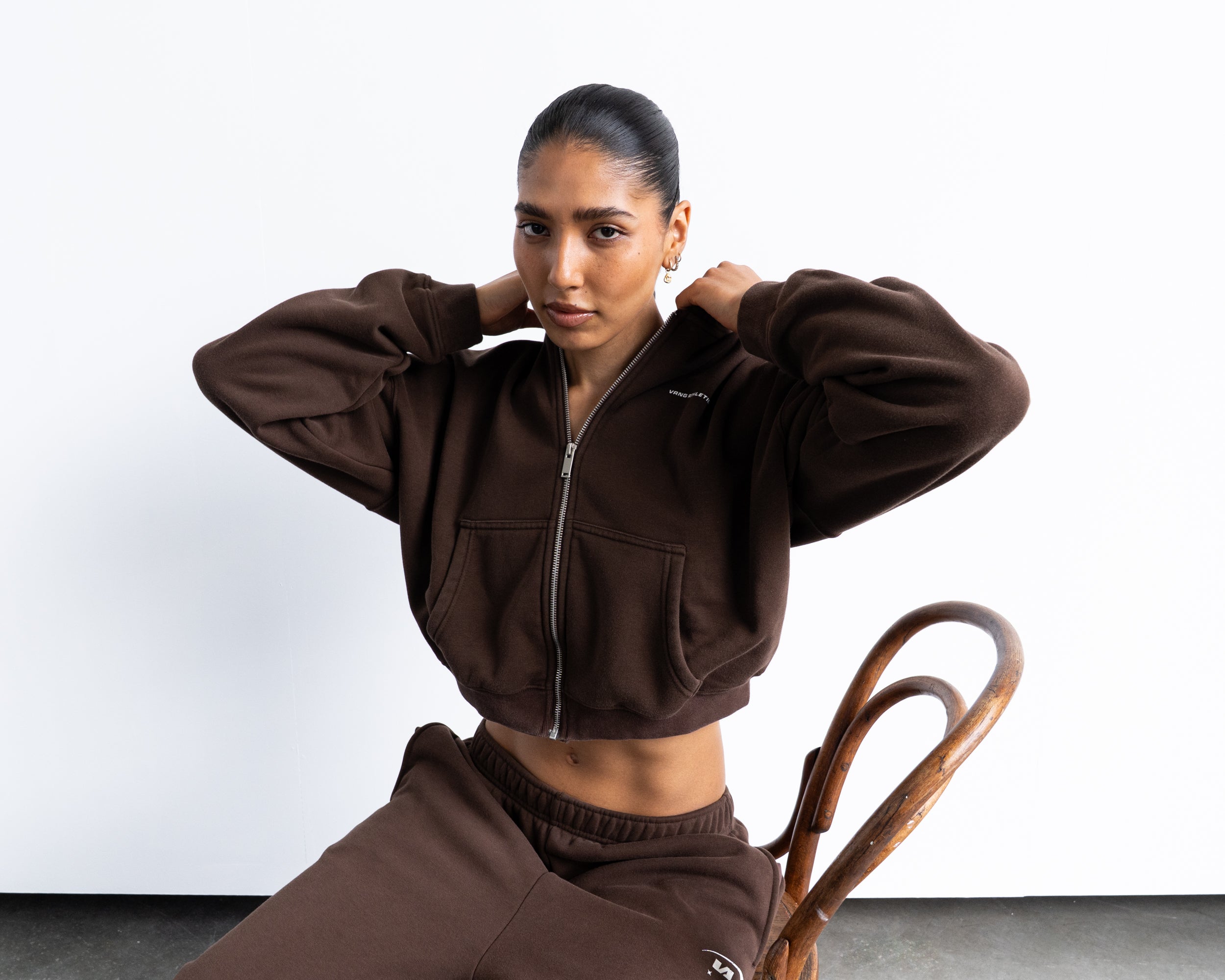 A model wearing a matching dark brown cropped zip-up hoodie and sweatpants set, sitting on a wooden chair against a plain white studio background.